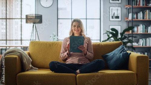 Beautiful Caucasian Female Using Tablet Computer in Stylish Living Room while Sitting on a Cozy Couch Sofa. Young Woman Resting at Home, Browsing Internet, Using Social Networks, Having Fun in Flat. photo