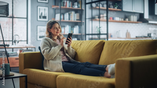 Beautiful Caucasian Female Using Smartphone in Stylish Living Room while Resting on a Cozy Couch Sofa. Young Woman at Home, Browsing Internet, Using Social Networks, Having Fun in Flat.