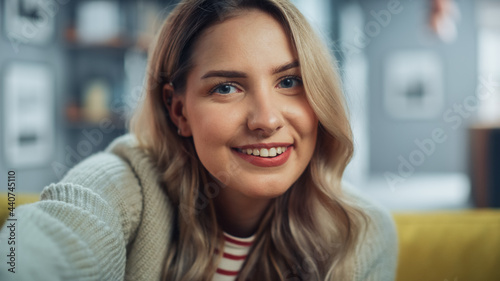Portrait of Beautiful Young Woman with Light Brown Hair Wearing White Woolen Sweater Looking Up to the Camera and Smiling Charmingly. Successful Woman Resting in Bright Living Room.