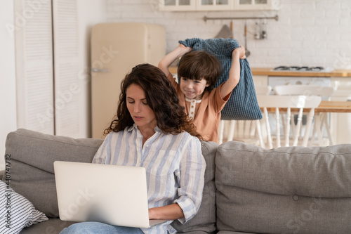 Foto Concentrated young woman use laptop computer with playful little kid hit her with pillow