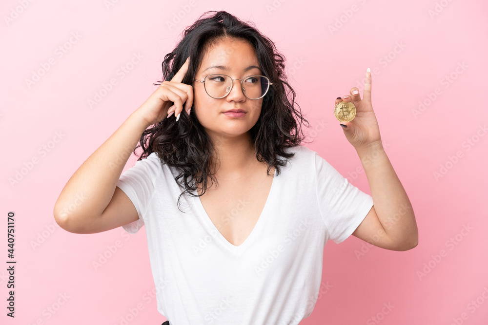Young asian woman holding a Bitcoin isolated on pink background having doubts and thinking Young asian woman holding a Bitcoin isolated on pink background having doubts and thinking
