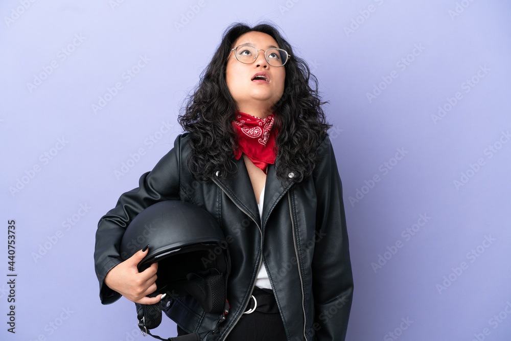 Young asian woman with a motorcycle helmet isolated on purple background looking up and with surprised expression Young asian woman with a motorcycle helmet isolated on purple background looking up and with surprised expression