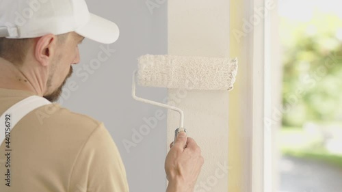 painter man decorator work of home to renovate, painting with roller paint on wall, window with green as background, close up