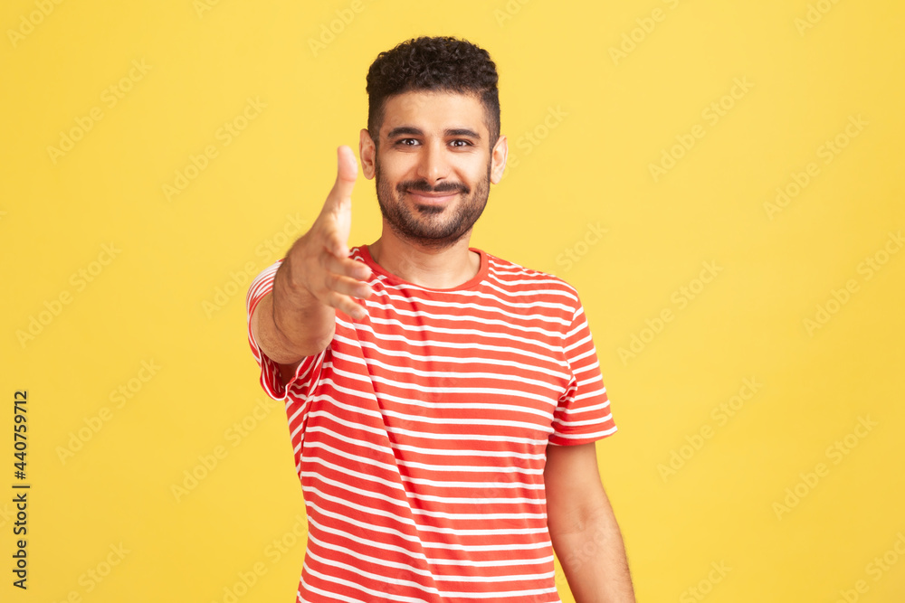 Nice to meet you. Positive friendly man with beard in striped t-shirt giving hand for handshaking, welcoming partners, job offer. Indoor studio shot isolated on yellow background Nice to meet you. Positive friendly man with beard in striped t-shirt giving hand for handshaking, welcoming partners, job offer. Indoor studio shot isolated on yellow background
