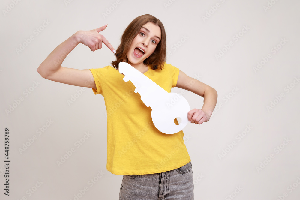 Portrait of funny positive teenager girl in casual style yellow T-shirt pointing at big paper key and looking at camera with toothy smile, purchase. Indoor studio shot isolated on gray background.