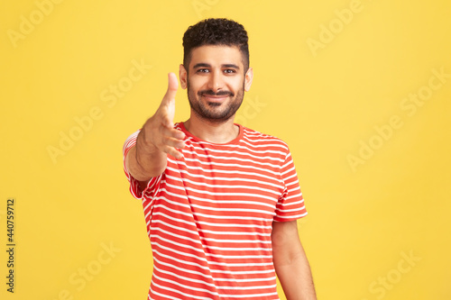Nice to meet you. Positive friendly man with beard in striped t-shirt giving hand for handshaking, welcoming partners, job offer. Indoor studio shot isolated on yellow background