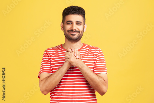 Happy positive man with beard in striped shirt pressing hands together on chest enjoying, calming and comforting himself. Indoor studio shot isolated on yellow background