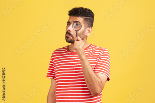 Serious bearded man in red striped t-shirt standing, holding magnifying glass and looking at camera with big zoom eye, verifying authenticity