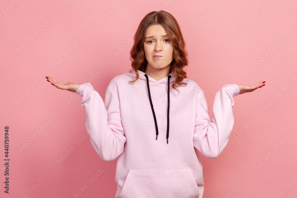 Portrait of confused curly haired teenager female in hoodie standing with raised arms and looking at camera with answer, dont know gesture. Indoor studio shot isolated on pink background Portrait of confused curly haired teenager female in hoodie standing with raised arms and looking at camera with answer, dont know gesture. Indoor studio shot isolated on pink background