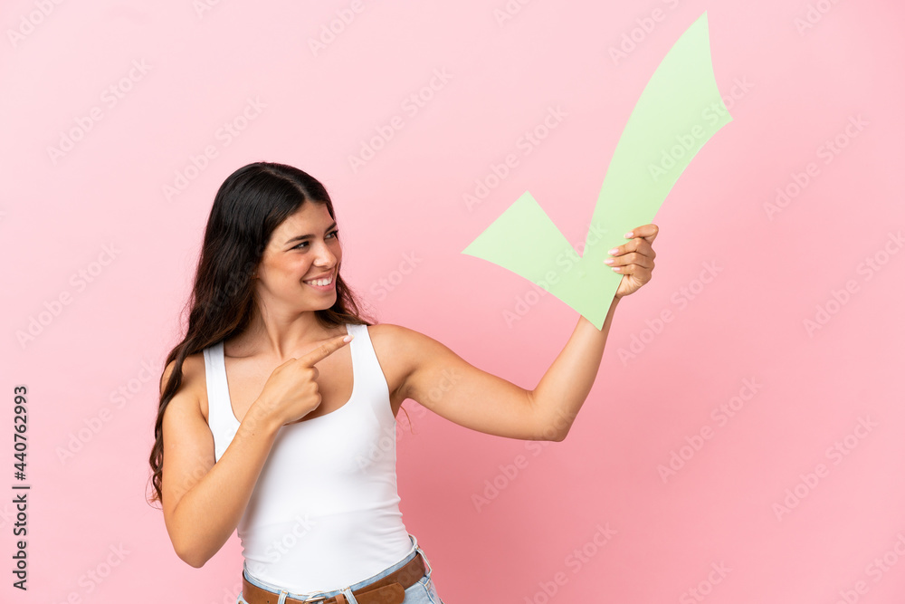 Young caucasian woman isolated on pink background holding a check icon and pointing it Young caucasian woman isolated on pink background holding a check icon and pointing it