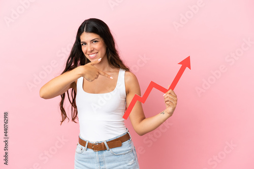 Young caucasian woman isolated on pink background holding a catching a rising arrow and pointing it