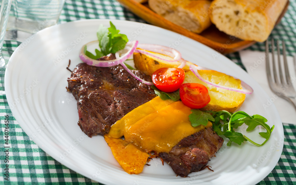 Appetizing well done roasted beef steak with cheese served with baked potato and fresh vegetables on white plate