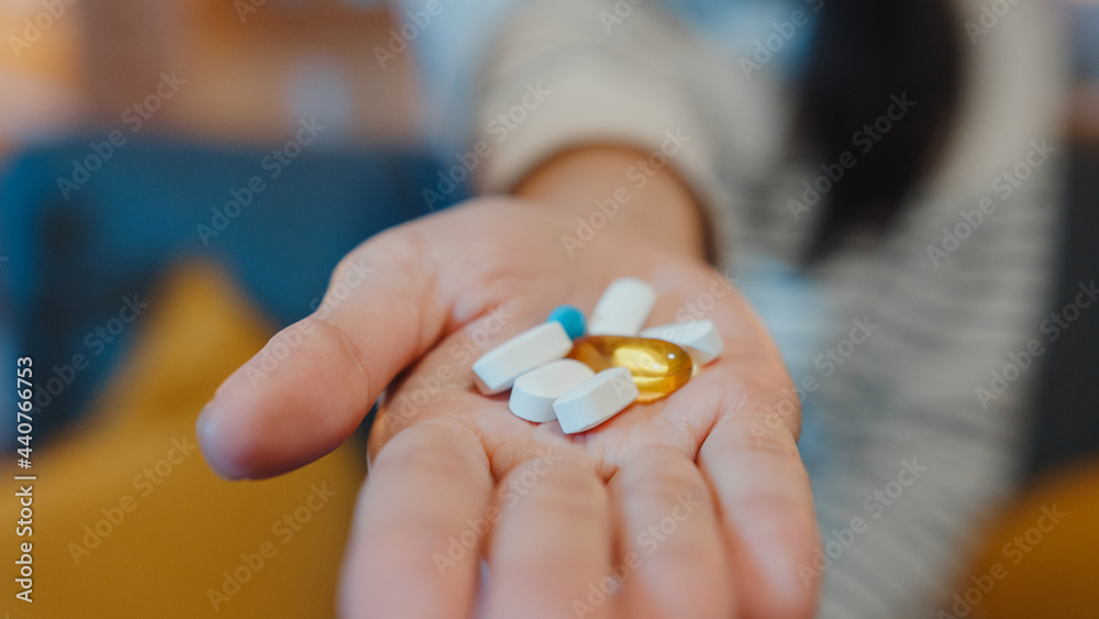 Sick asian young lady holding pill take medicine sit on couch in living room at home night. Girl taking drug after doctor order, quarantine at house, Coronavirus social distancing healthcare concept.