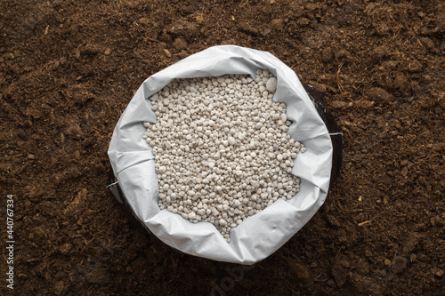 Opened plastic bag with gray complex fertiliser granules on dark soil background. Closeup. Product for root feeding of vegetables, flowers and plants. Top down view. photo