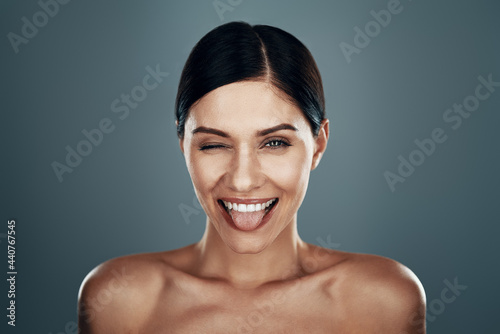 Playful young woman sticking out tongue and smiling while standing against grey background