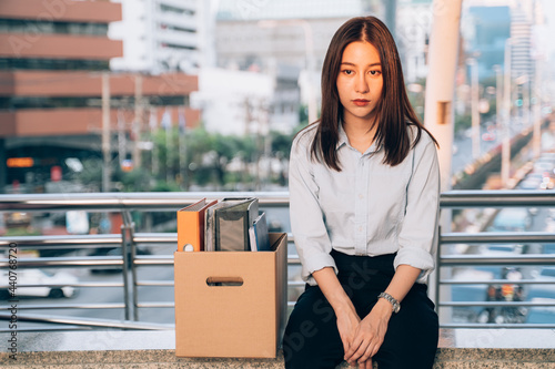 Stressed and worried young Asian woman with box of items sitting alone after being laid off from job due to recession and economic stress in industry photo