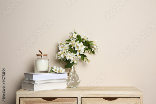 Bouquet of beautiful jasmine flowers in glass vase, books and candle on wooden commode near beige wall indoors