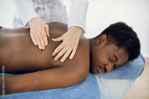Close up of young African-American man getting back massage. Hands of european woman doctor chiropractor making massage for back pain relief for young guy.