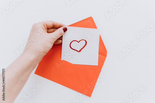 A woman's hand puts a drawing of a red heart in an envelope.On a white background.Top view.Love, friendship, and a declaration of love.Happy Valentine's Day.Women's Day.Minimal holiday concept