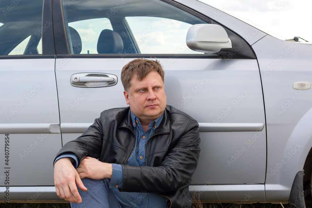 A tired driver sits on the grass near the car in the steppe. A tired driver sits on the grass near the car in the steppe.