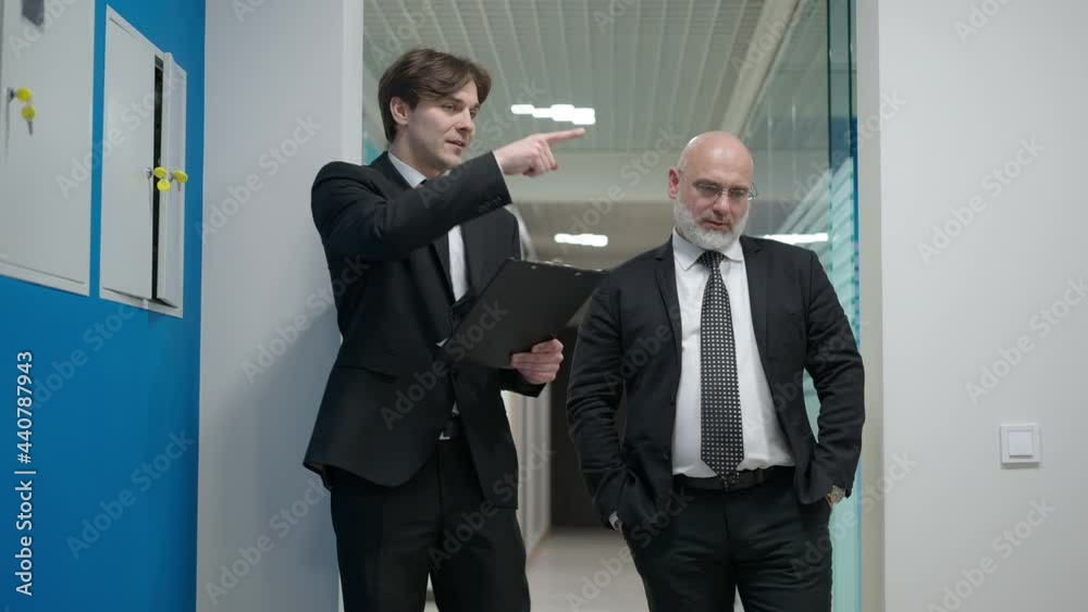 Medium shot front view of two confident businessmen talking walking in hallway indoors. Portrait of young and middle aged Caucasian men discussing business project strolling in office corridor Medium shot front view of two confident businessmen talking walking in hallway indoors. Portrait of young and middle aged Caucasian men discussing business project strolling in office corridor