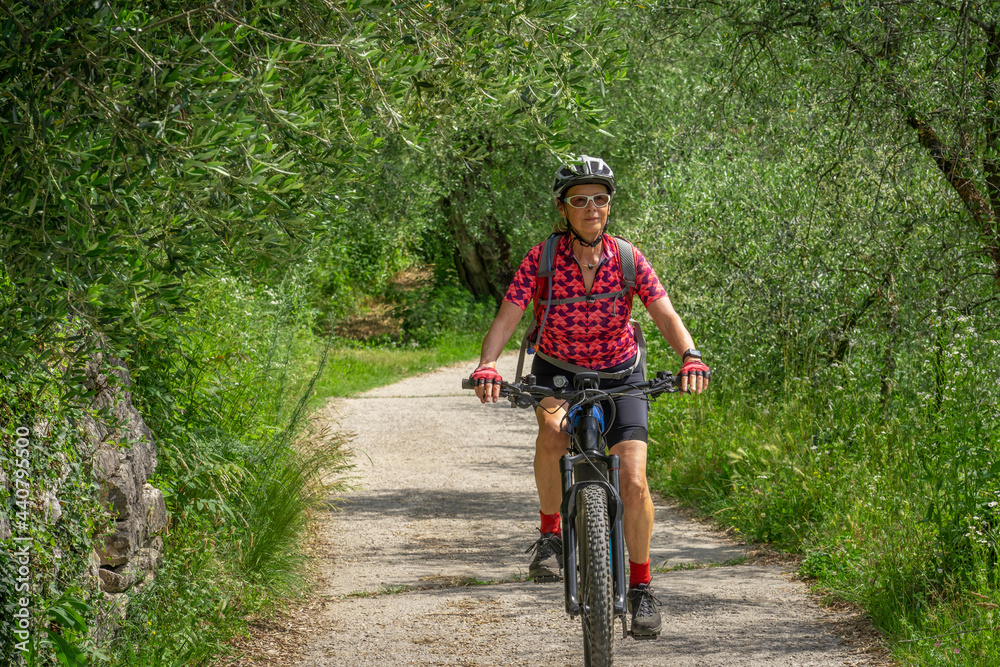 Fototapeta premium nice and active senior woman riding her electric mountain bike in the Garda lake mountains between blooming the olive groves of Arco close to Riva del Garda and Garda Lake , Landscape