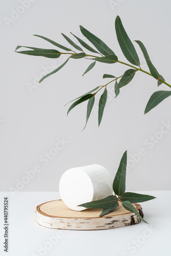 White container on wooden podium and eucalyptus branch on grey background. Round jar for natural cosmetic product branding on birch saw cut and green leaves. Beauty and spa advertising mockup.