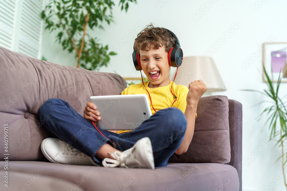 happy boy with headphones and a tablet is sitting at home on couch. child is happy to learn at home online on computer tablet, listening to music. new children's learning app. Dependence on gadgets happy boy with headphones and a tablet is sitting at home on couch. child is happy to learn at home online on computer tablet, listening to music. new children's learning app. Dependence on gadgets