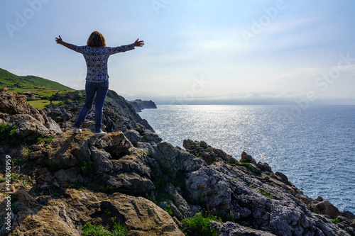 Woman climbing a stone with open arms and looking at the sea from the cliffs.