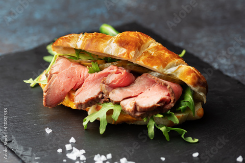 Fresh baked croissant sandwich with arugula and roast beef on a black slate board