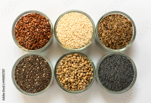 An assortment of healthy seeds (chia, hemp flax, dill, sesame, poppy seeds) - top view of small glass bowls on a white background.
