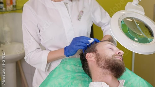 Mesotherapy treatment for hair. Female doctor in rubber gloves preparing man head and hair before injecting vitamins into scalp, wiping it with cotton pad while working in cosmetology beauty clinic photo