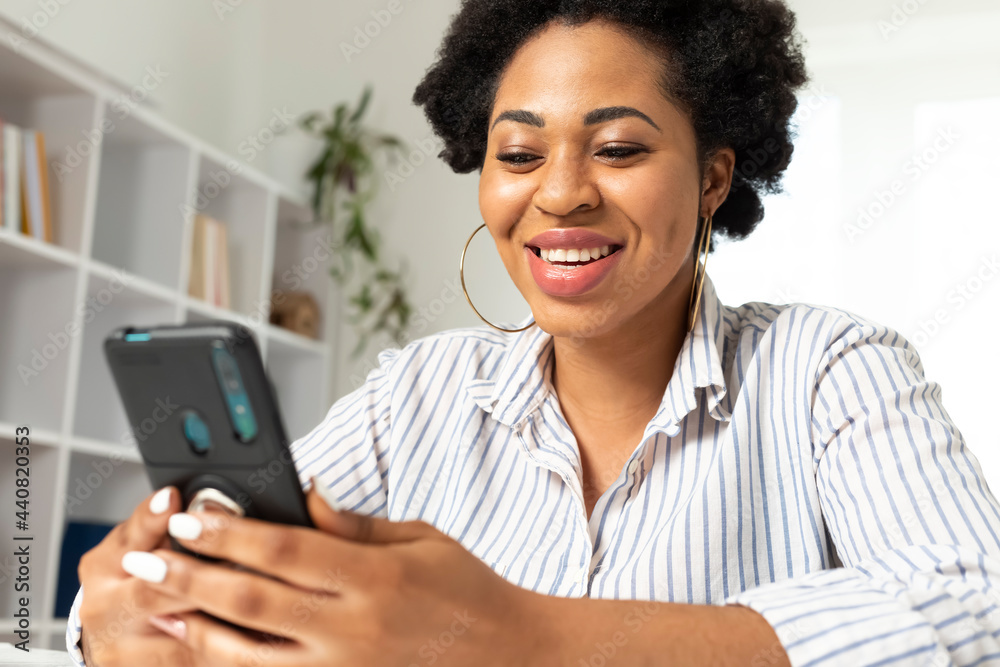 Portrait of a happy African American woman using mobile phone at home. Black smiling female chatting holding a smartphone in her hand in a light interior. Portrait of a happy African American woman using mobile phone at home. Black smiling female chatting holding a smartphone in her hand in a light interior.
