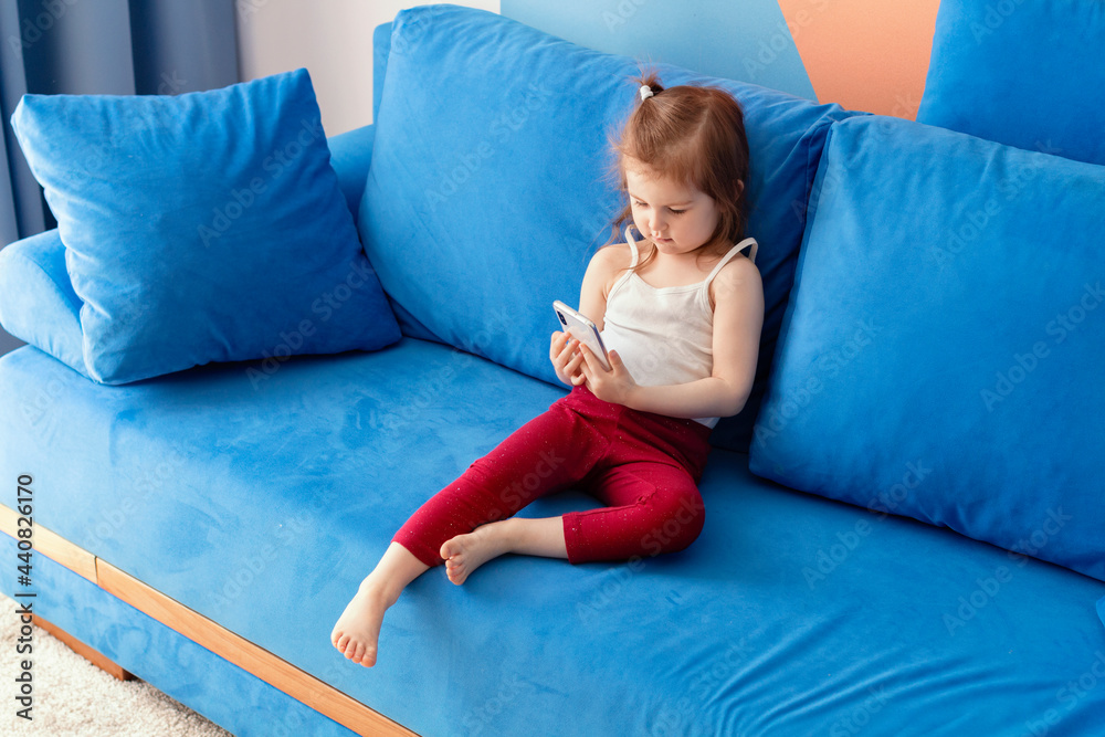 Cute little toddler girl wear casual clothes sitting on blue coach in domestic room and using smartphone. Cute little toddler girl wear casual clothes sitting on blue coach in domestic room and using smartphone.