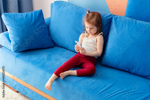 Cute little toddler girl wear casual clothes sitting on blue coach in domestic room and using smartphone.