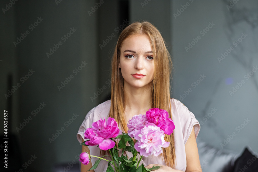 A beautiful girl with long blonde hair in a modern interior holds a bouquet of pink peonies in her hands, a romantic girl