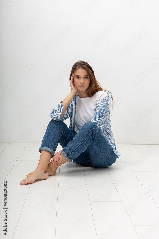 portrait of young attractive caucasian woman with long brown hair in t-shirt and blue jeans isolated on white background. skinny pretty female sitting on floor at studio. model tests of beautiful lady