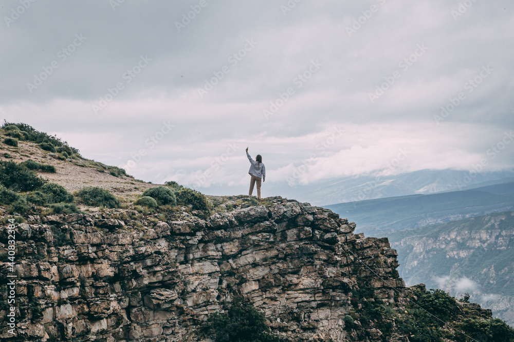 A young traveling woman stands on the edge of a mountain cliff and watching a beautiful view of the forest, mountains and clouds on vacation. The woman travels alone. Dagestan mountains