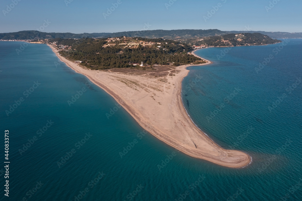Aerial view to a long narrow sandy beach stretched into the turquoise sea with a white lighthouse, dense forest, and small town on a hill in the background