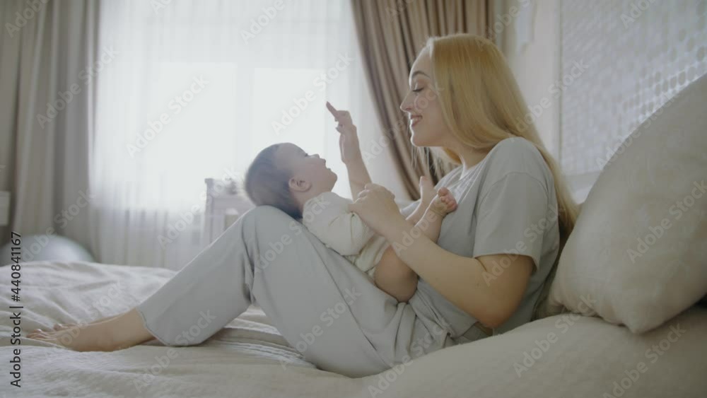 Mom plays with her newborn baby at home. Portrait of young mother and his son sitting on bed