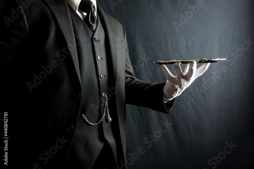 Portrait of Butler in Dark Suit and White Gloves Beautifully Holding Silver Serving Tray on Black Background. Concept of Service Industry and Professional Hospitality. 