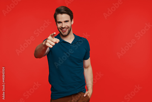 Happy young man in casual wear pointing camera while standing against red background