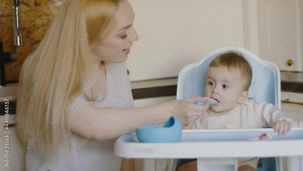 Mom feeds little baby boy with spoon. Small child eats at home while sitting in highchair Mom feeds little baby boy with spoon. Small child eats at home while sitting in highchair