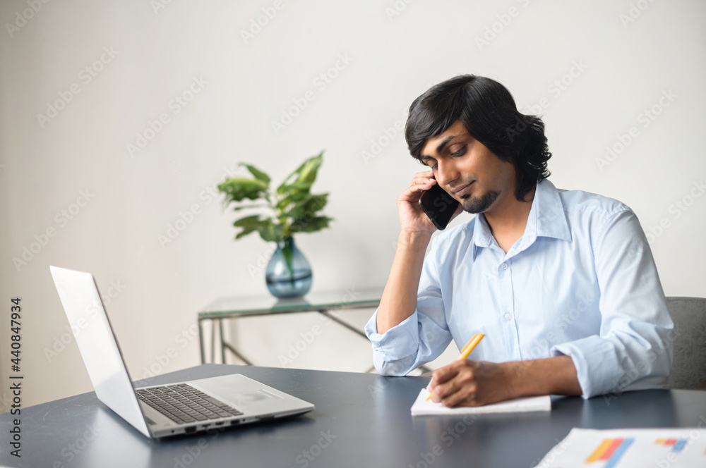Intelligent indian male office employee talking on the smartphone and taking notes sitting at the desk in bright office space, using laptop computer, eastern man writes down during phone conversation Intelligent indian male office employee talking on the smartphone and taking notes sitting at the desk in bright office space, using laptop computer, eastern man writes down during phone conversation