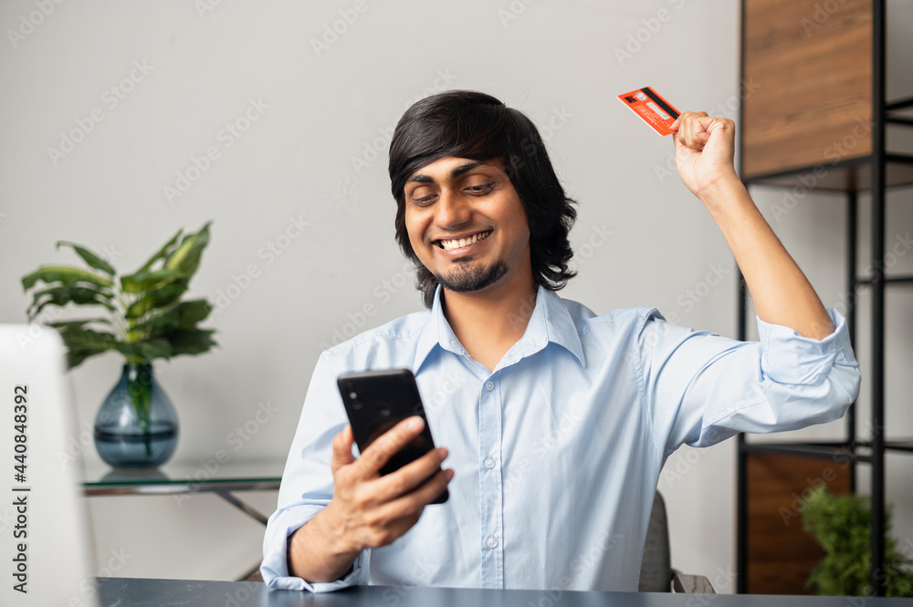 Happy indian man holds banking card and smartphone, made long awaited purchase and celebrates it, buying and shopping online, using mobile app for e-shopping Happy indian man holds banking card and smartphone, made long awaited purchase and celebrates it, buying and shopping online, using mobile app for e-shopping