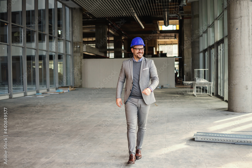 Smiling businessman with helmet on head walking trough construction site of his building.