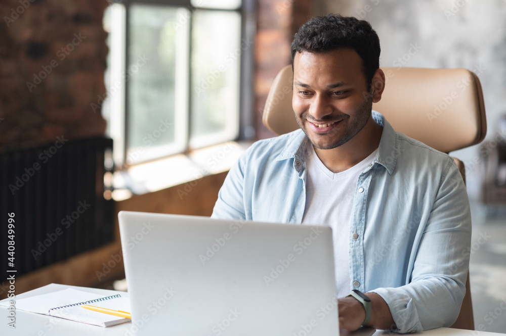 Intelligent indian businessman using a laptop in the office. Smart smiling man entrepreneur looks through emails, analyzes tasks, checks report. Hispanic freelancer in modern loft office Intelligent indian businessman using a laptop in the office. Smart smiling man entrepreneur looks through emails, analyzes tasks, checks report. Hispanic freelancer in modern loft office