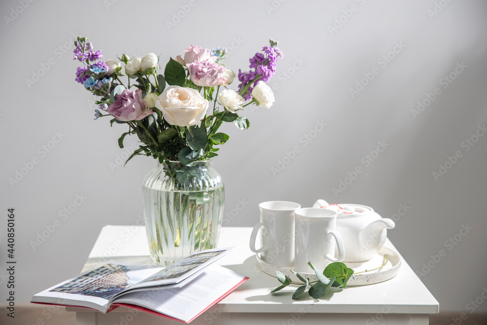 Bouquet of hackelia velutina, purple and white roses, small tea roses, matthiola incana and blue iris in glass vase is on the white coffee table with two tall cups for tea. Grey wall