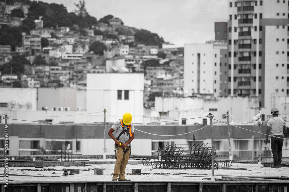 on top of a construction site using safety equipment. Helmet, seat belt and reflective vest. In the background a slum in black and white on top of a construction site using safety equipment. Helmet, seat belt and reflective vest. In the background a slum in black and white