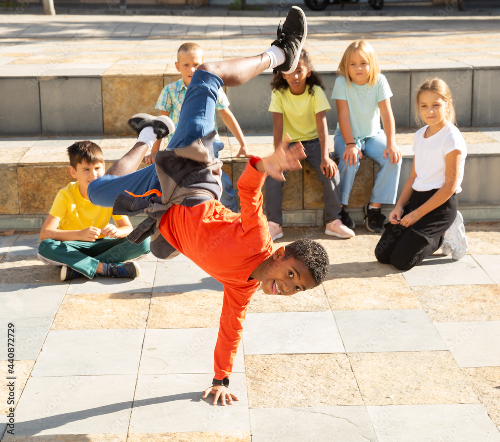 Cheerful b-boy performing stunts on city street, doing hand hop in front of group of teens
